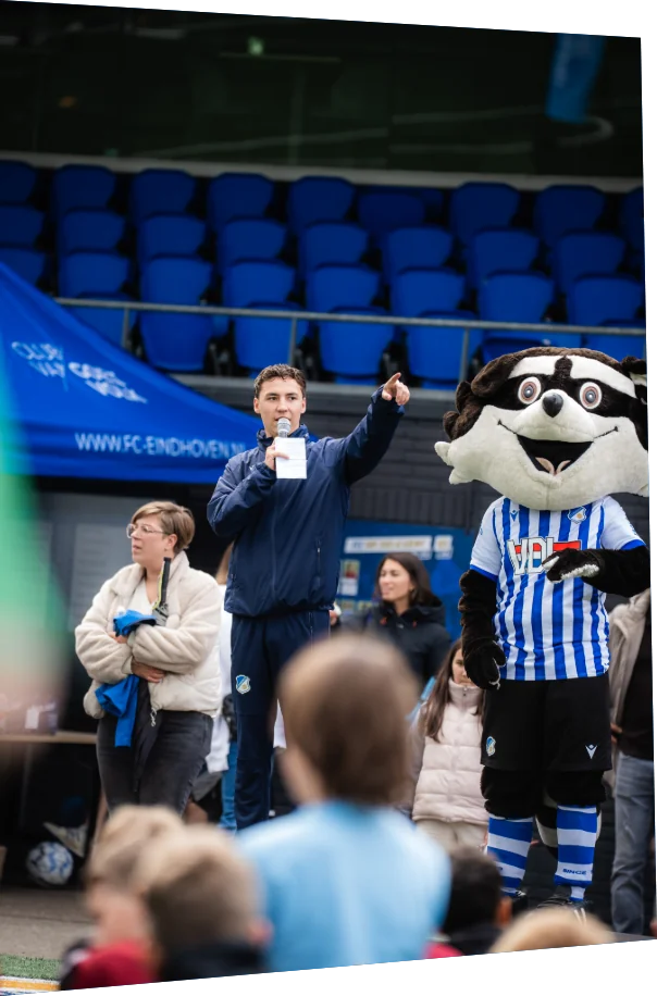 Presentator met microfoon en de mascotte van FC Eindhoven spreken de schoolteams toe tijdens het ASML Schoolvoetbaltoernooi
